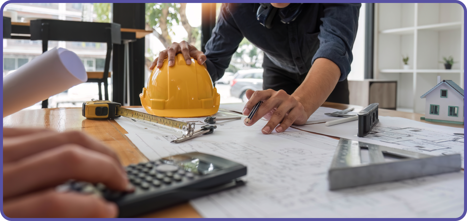 Man with hardhat in hand leaning over a table with architect's drawings and calculator on table
