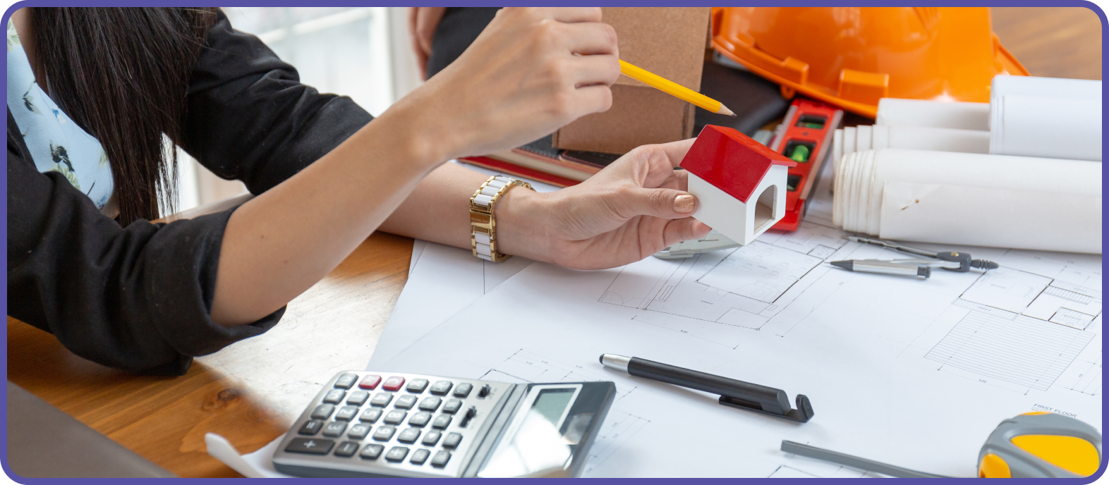 Woman with a small model of a house in one hand, and a pencil in the other hand sitting at a table with drawings, calculator and pen on the table.
